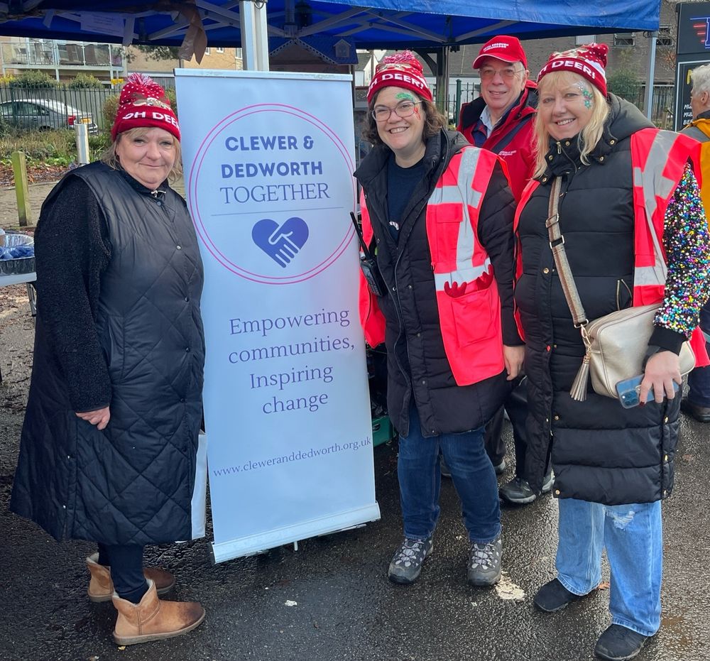 Lee, Amy and Alison, wearing festive bobble hats, stand in front of a sign saying Clewer and Dedworth Together. Empowering Communities, Inspiring Change. An RBWM Ambassador is photobombing in the background! 
