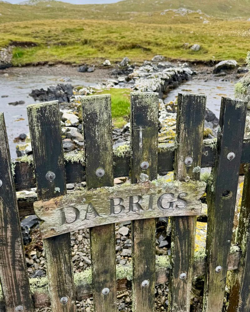 Upper section of an old and very weathered gate. It is made from vertical wooden slats bolted onto horizontal slats. The vertical slats are positioned so that they make a pleasing curve at the top. There is a curved wooden sign attached part way down which says DA BRIGS. Beyond the gate is a narrow stony causeway over some water leading to a rough grassy area. A glimpse of grey sky beyond.