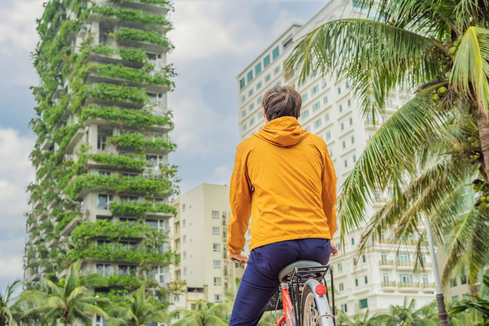 A cyclist stands in front of a tall residential building with plants growing from its balconies
