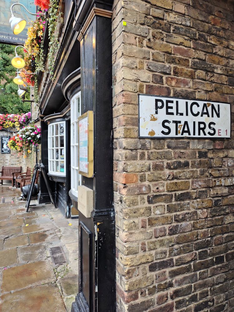 Street sign on the side of The Prospect of Whitby pub in Wapping, London reads "Pelican Stairs." The front of the pub is festooned with hanging baskets of flowers.