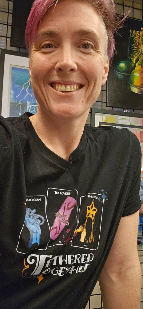 Smiling person in their '40s, wearing a black T-shirt that says tethered together and has three tarot cards on it. The cards are the magician, the lovers, and the star.