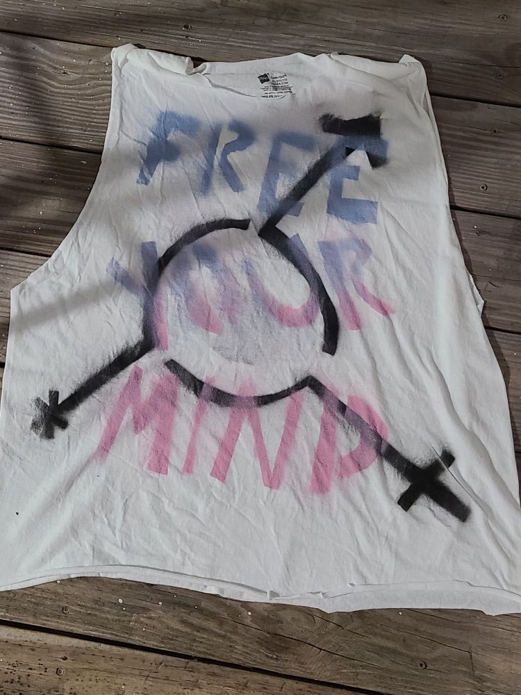 A photo of a handmade tank top that reads FREE YOUR MIND in transgender pride colours, laid over a black transgender symbol. 