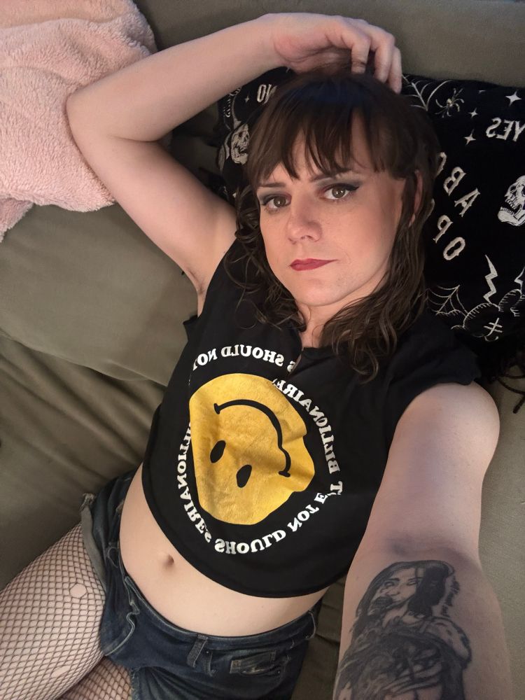 Izzy lying back taking a selfie with one harm resting on the couch above her. She’s wearing fishnets, cutoff jean shorts, and a crop top with an upside down smiley and the words billionaires should not exist