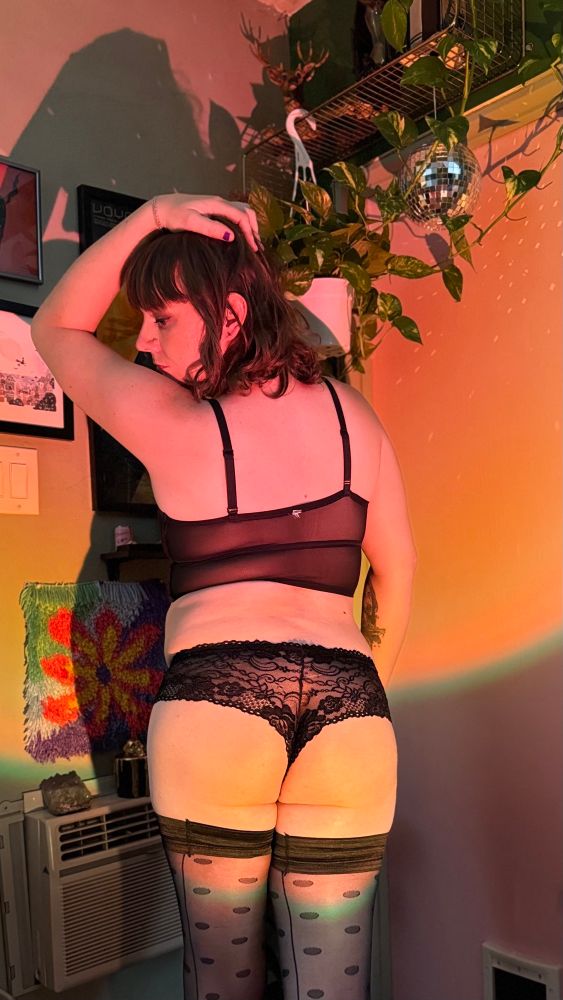Izzy from behind, standing in an orange and yellow light, looking over her shoulder thru the window created by her lifted arm with hand resting on her head. She’s wearing a sheer black top, lacy black undies, and thigh high nylons 