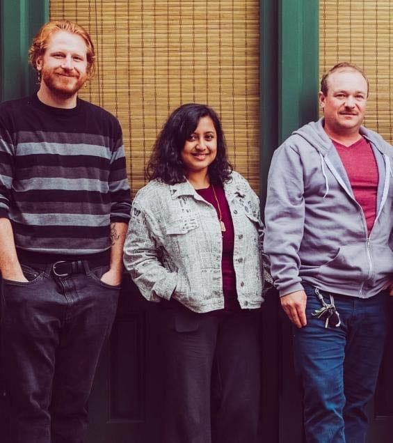 Left to right: man leaning against wall with hands in jeans pockets, woman leaning against wall with hands in jacket pockets, man leaning against wall 