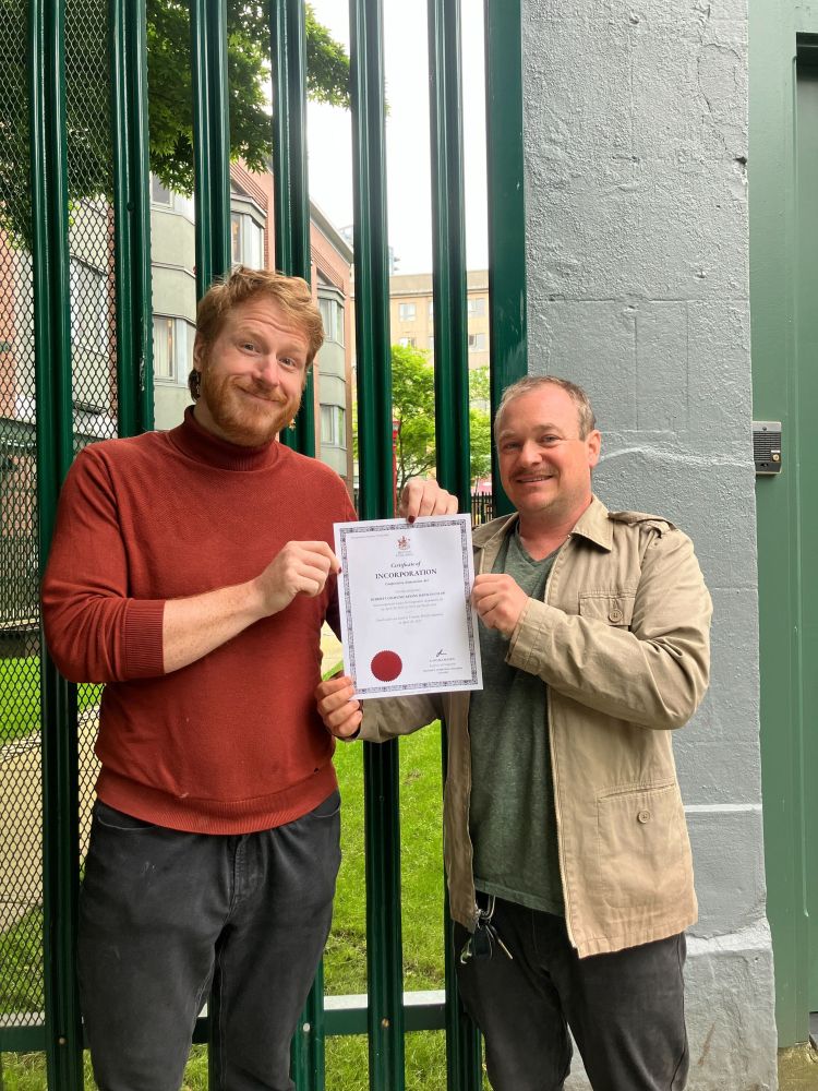 Two people standing in front of a green gate, holding a co-op business registration certificate.