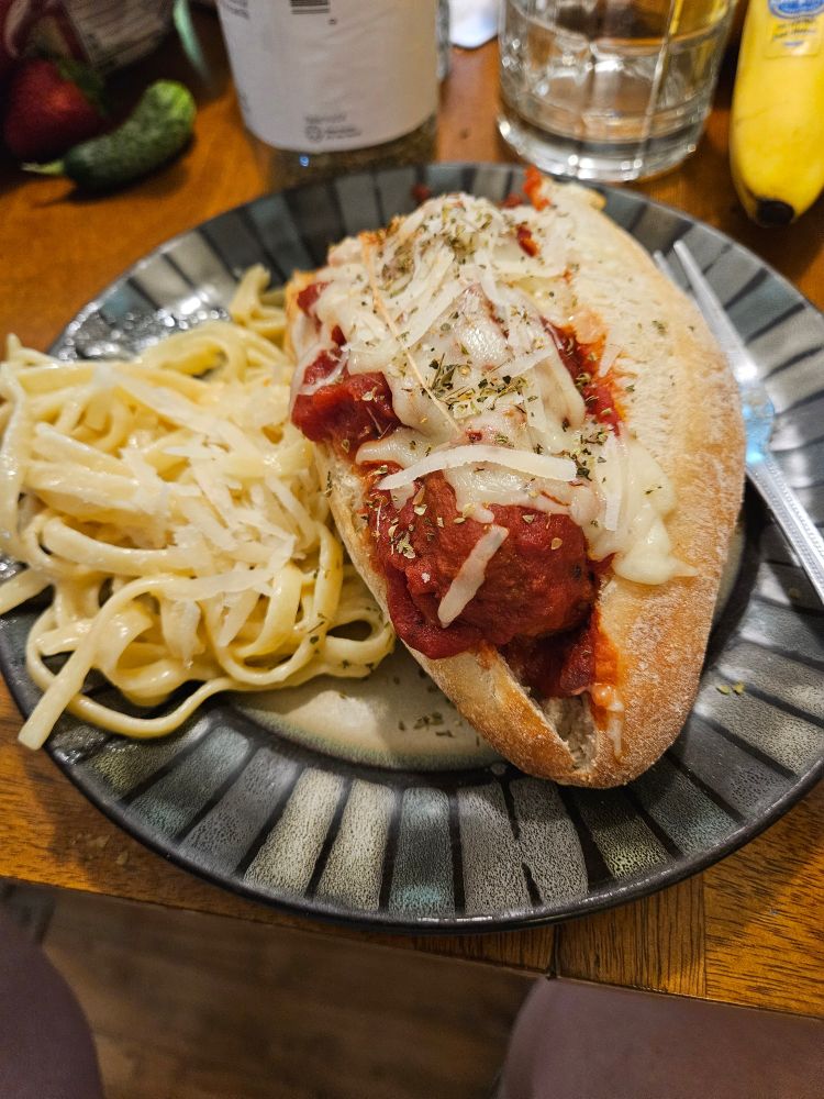 Meatball sandwich and fettucine alfredo with parmesan on a dark blue and gray plate. There is, from left, a small green cucumber, a container of oregano, a small glass, and a banana behind the plate.
