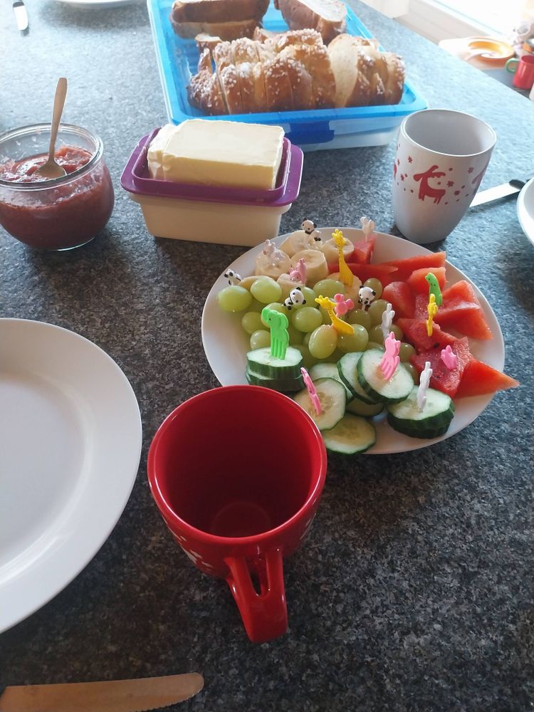 Der Frühstückstisch, im Vordergrund ein Teller mit Gurke,Trauben, Bananen Melone, dahinter ein Stück Butter, ein Glas Marmelade mit Löffel, Hefezopf, im Vordergrund eine rote und eine weiße Tasse jeweils mit Elchen drauf
