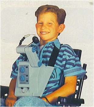  A smiling boy sits in a chair wearing a blue striped shirt and a large, Nintendo Hands Free Controller strapped to his chest. 