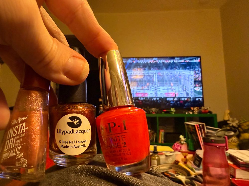 Three nail polishes, two copper colors and a reddish orange. All three of those in front of the TV showing a wrestling cage match