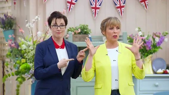 A screencap from The Great British Baking Show, of Sue and Mel. Sue is pointing at Mel, who is gesturing and talking to the bakers about their next challenge.
