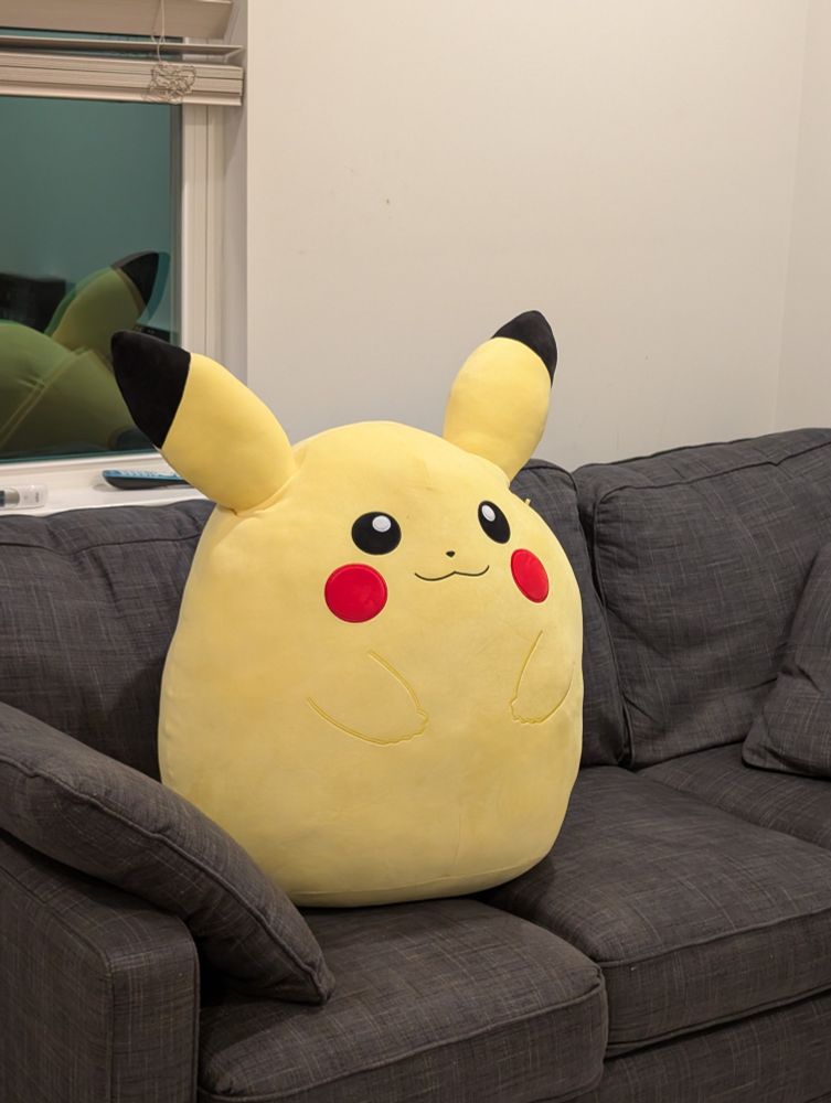 A giant, oval Squishmallow Pikachu plush sitting on a full-sized couch. It takes the spot of an entire adult.