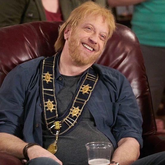 A picture of Roland Schitt from Schitt's Creek. He's a ginger white man with a beard, wearing a grey shirt and open dark navy button down shirt with a medal draped over his neck. He's sitting in a dark red leather chair and smiling as he holds a clear plastic cup with beer in it.