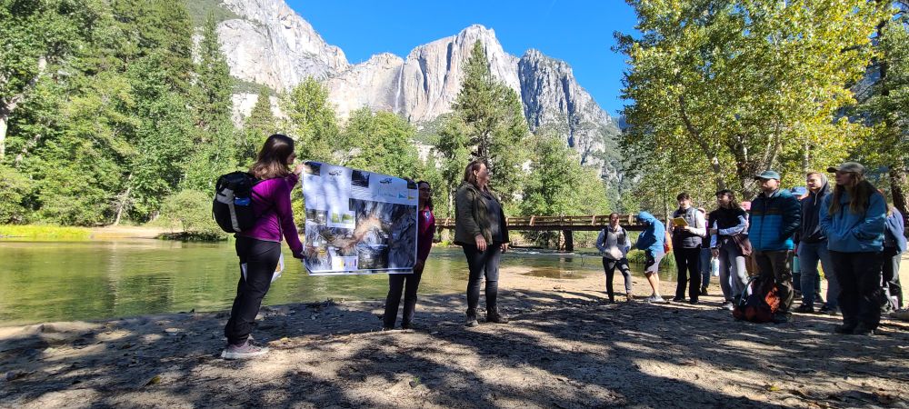 Group of people are gathered along the sandy bank of the Merced River. Two volunteers are holding up a poster while the presenter speaks to the rest of the group. Behind the poster and presenter is Yosemite Falls. 