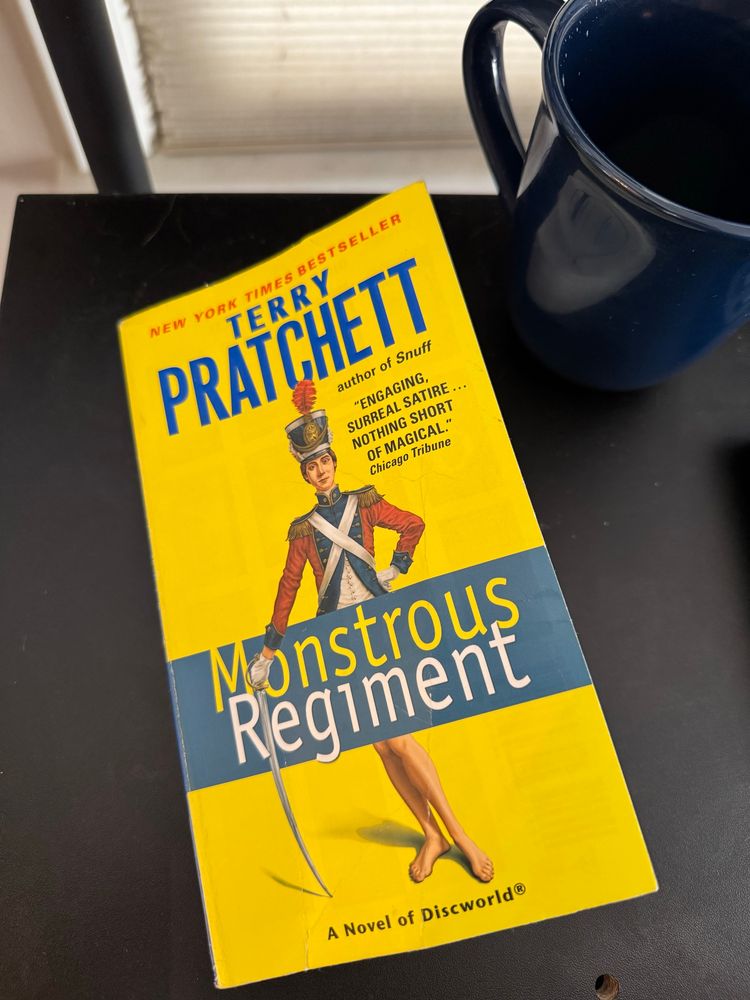 Cover of the book “monstrous regiment” by terry prachett. Book is yellow and depicts a solder in a red and blue uniform posing with a sword. From the waist down is a set of bare legs, with the waist covered by the text “Monstrous Regiment”.  Top of the book reads “New York times bestseller terry prachett”
