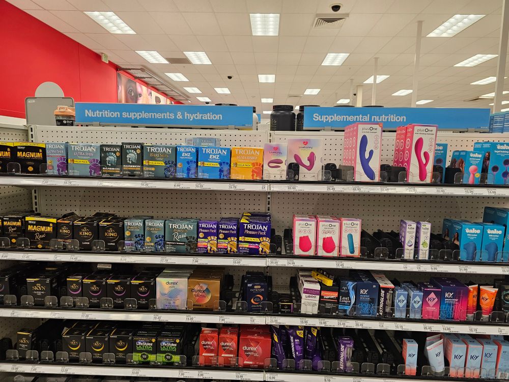 Picture of semi-stocked shelving from a Target. To the left the top is labeled "Nutrition Supplements & Hydration." The shelves below it are filled with an assortment of condoms. To the right, the top is labeled "Supplemental Nutrition." The shelves below it are filled with women-targeted sex toys on the top 2 shelves and various lubes and other products for intercourse.