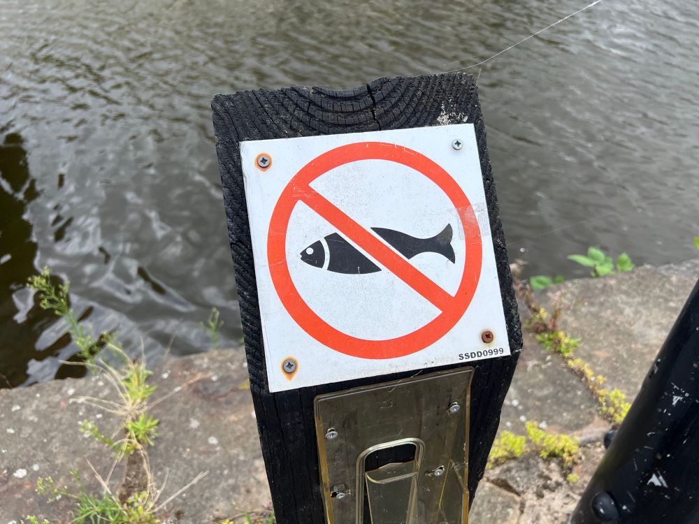 A sign by a canal showing a fish with a line through it. Presumably saying no fishing, but it looks like no fish. 