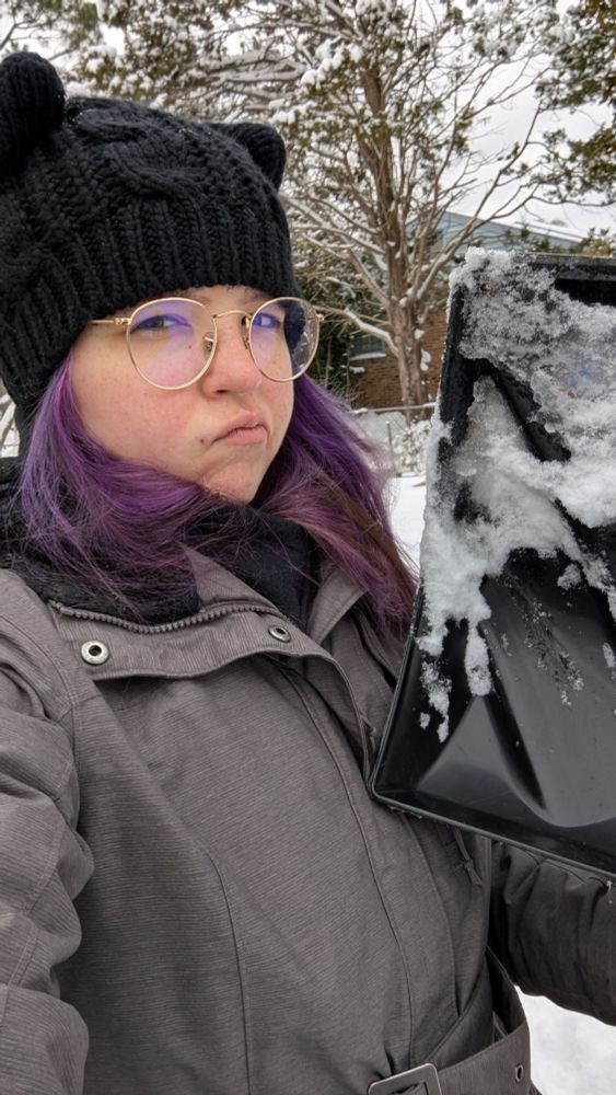 a selfie of a woman with purple hair and glasses wearing winter gear. she is holding a snow shovel and making a determined expression at the camera.