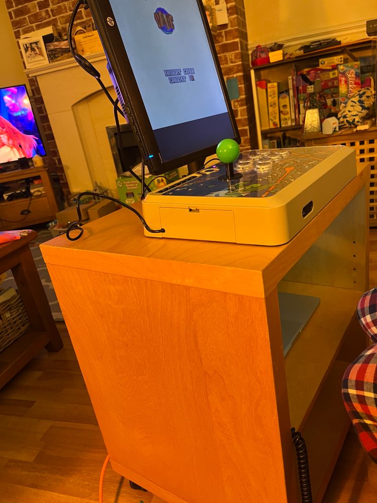 ´Mobile Arcade’ or ´Dad Cab’ setup. 2013 Apple MacBook attached to a monitor and arcade stick on a rolling two tier small shelving unit. The game displayed is Dodonpachi Saidaioujou (Knuckles version). Strictly is on on the TV in the background.