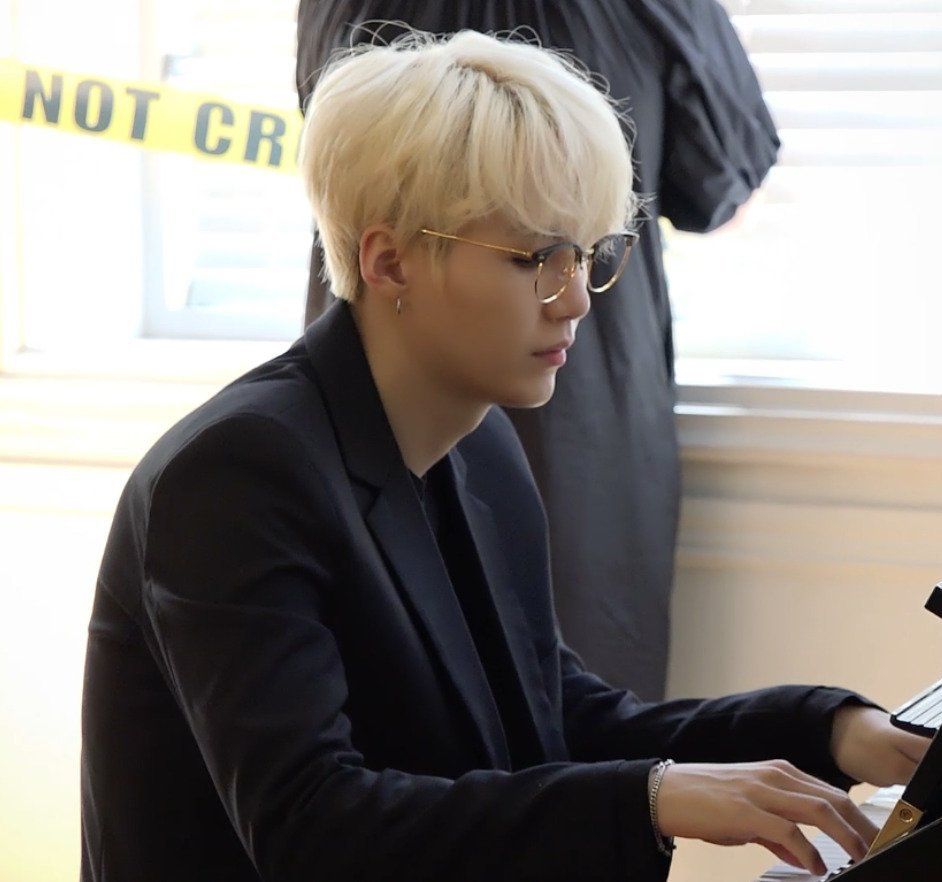 Blond Yoongi wearing a black jacket over a black shirt and good rimmed glasses playing a piano. In the background there is the back of a figure in black next to some yellow "Do not cross" tape.