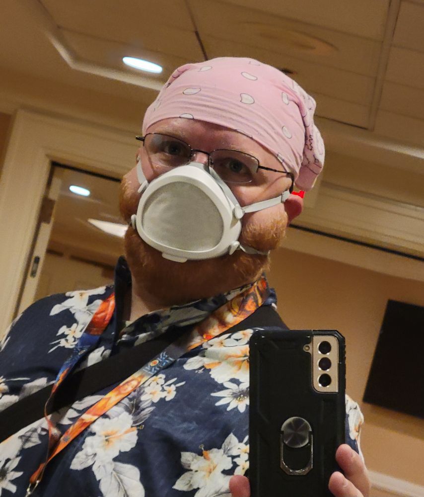 A white enby with a beard, floral shirt, pink bandana and Flo mask