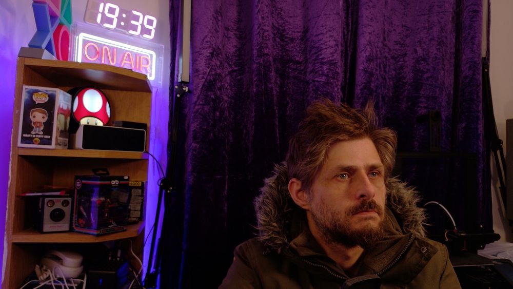 Me sat in a thick green outdoor coat in front of a pair of closed purple velvet curtains. Behind my right shoulder is a wooden corner shelf, rear-illuminated in purple. On the top shelf is a large RGB clock (displaying the time 19:39) on top of a neon-style LED 'on air' light (text in red surrounded by a white rounded rectangle, embedded in clear plastic), next to a PlayStation light (not turned on, comprising the iconic square, triangle, cross and circle in a two-by-two grid). On the shelf below is a Marty McFly funko pop, an illuminated super mushroom from Super Mario, and a Ulanzi and Divoom LED matrix (both turned off). The next shelf has a Kodak Box Brownie camera and Casio organiser. A white landline phone is visible below those.