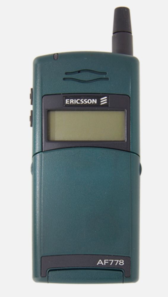 A view of a dark green Erikson cell phone from the late 1990s. It is rectangular. It has a closed cover over the keypad that is openable. The phone's antenna on the right is short and stubby and colored black. 

The phone screen is very small and the background is green. Above the screen is the Erickson brand. At the bottom right of the phone is the model: AF778.