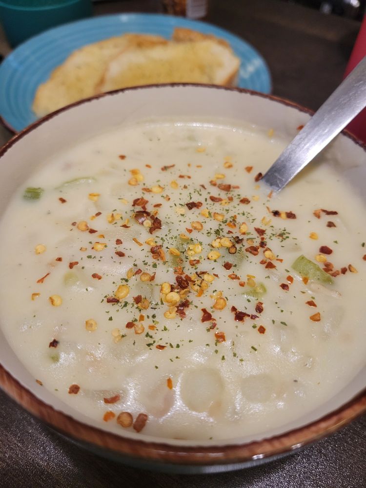 The same bowl of chowder as the second photo, white topped with red pepper flakes and dried green parsley. In the background, a blue plate holds two slices of toasted sourdough (oval) with butter and a sprinkle of dried parsley.