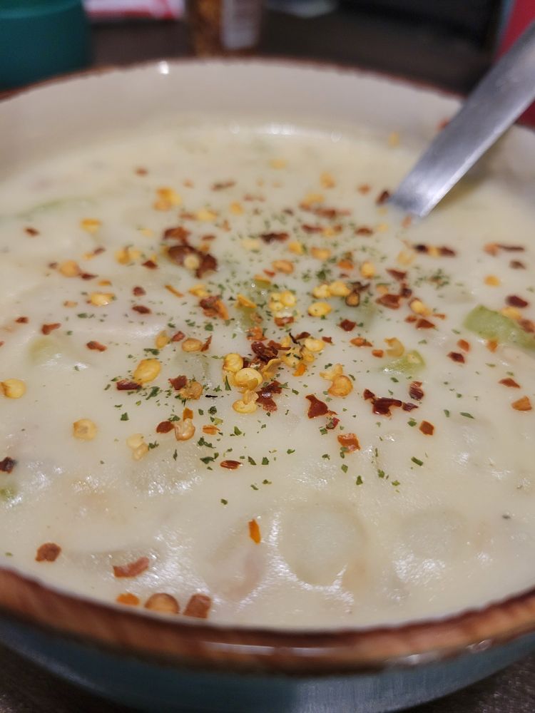 Close-up shot of clam chowder in a bowl with a brown rim. Sprinkled on top of the creamy chowder is red pepper flakes and a sprinkle of dried parsley. Potato and celery are visible in the chowder.