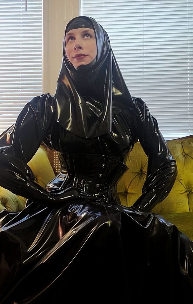 Woman sitting on a green couch wearing a large latex skirt, corset, loose fitting blouse, open faced hood and headscarf.