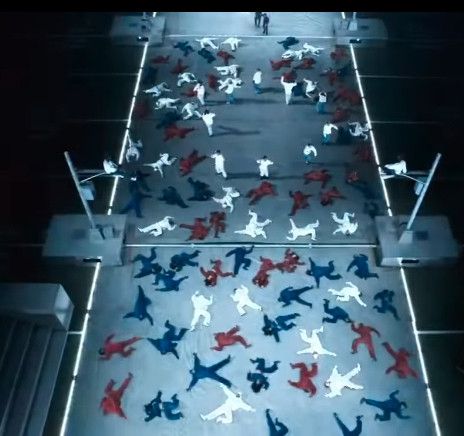 A bird's eye view still frame from Kendrick Lamar's Superbowl halftime performance, showing a bleak, gray urban street scene with industrial-looking surveillance cameras mounted on poles.

On the street, dozens of performers wearing red, white, and blue outfits are splayed out on the ground like corpses (almost looking like puzzle pieces or bodies ready for chalk outlines), while a small circle of performers wearing white are arranged in a small circle toward the top of the frame.