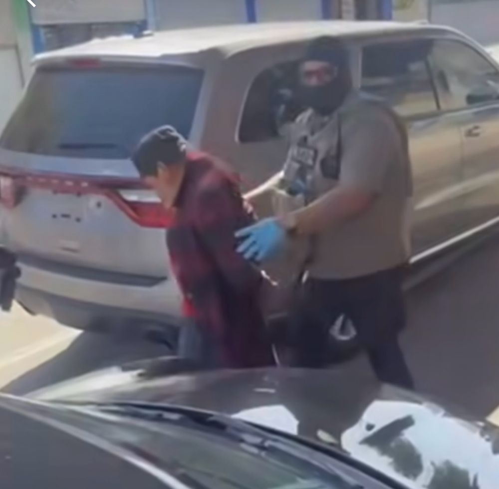 A large masked man with a bulletproof vest and blue nitrile gloves hustles an elderly man whose hands are cuffed behind his back into an unmarked vehicle with no rear plate