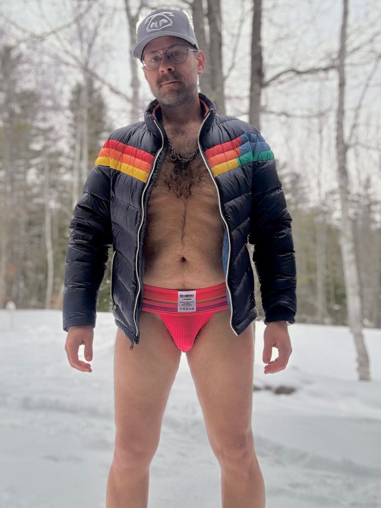 A man outside in a snowy forest, facing the camera, wearing a ball cap, a puffer jacket, and a neon pink jockstrap.