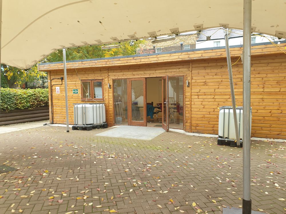 A low wooden cabin with a paved area in front, under a permanent tent like awning. It's surrounded by greenery.