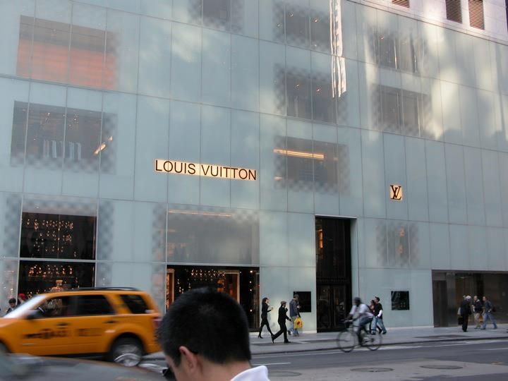 Louis Vuitton facade, Fifth Avenue, New York City (unveiled in 2004), in semi-opaque laminated glass with the designer's name and logo in the center, view of the street below with pedestrians, a cyclist, and a yellow taxi cab exiting the screen on the left; back of a person's head with dark short hair in the foreground.