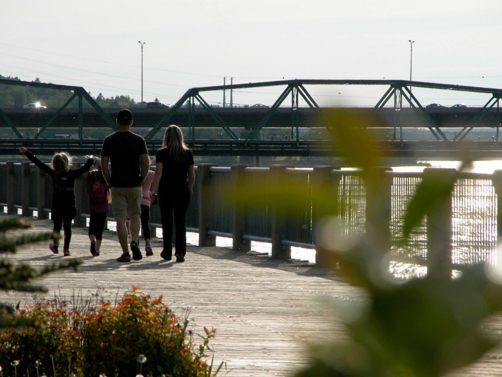 Family with a couple and children walking along the boardwalk on the riverfront with silhouette of Pont Sainte-Anne in the background at sunset