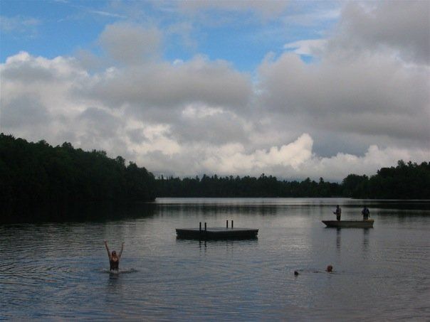 Lake view against cloudy blue sky with a raft in the centre, a swimmer jumping out of the water on the left and two people standing fishing on a rowboat on the right
