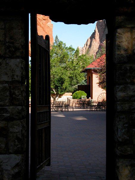 Narrow rectangular view from a dark old stone shed onto a sun-drenched courtyard with iron chairs and tables, red-roofed building, trees, sandstone formations, and blue sky in the background