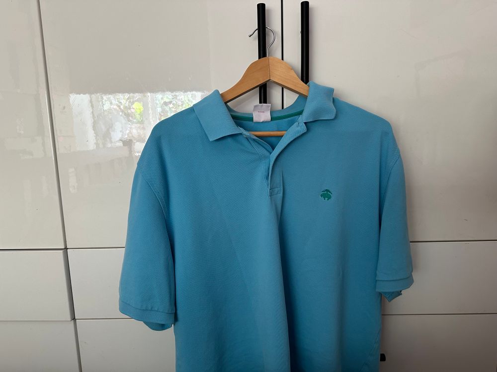 A polo shirt unbuttoned to the second button, on a birch wooden hanger, hanging on the black handle of a glossy white wardrobe cupboard door.  The shirt is a bright-ish mid blue. A small turquoise emblem on the breastplate reveals it comes from Brooks Brothers #BrooksBrothers 