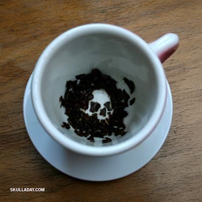 A white teacup viewed from above which shows a skull in the negative space of the black tea leaves at the bottom. The photo is marked skulladay.com as a source