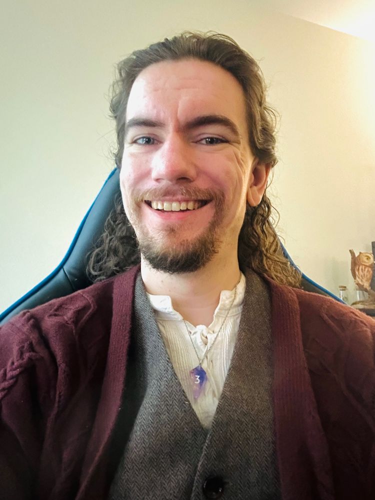 Paul, a nonbinary person, sits in a chair and grins at the camera. They have wavy shoulder-length hair and a short beard, and are wearing a brown vest and sweater, a white shirt, and a purple dice necklace