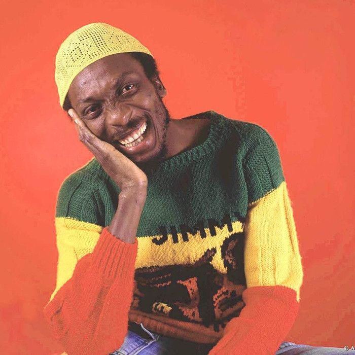 A young Jimmy Cliff in red gold and green sweater and wearing a yellow skull cap. He is smiling brightly and looks very happy. 