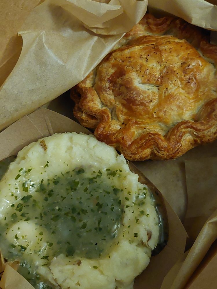 A small pie with a perfectly browned crust and a scoop of mashed potatoes with a green herb gravy in a brown paper takeout box