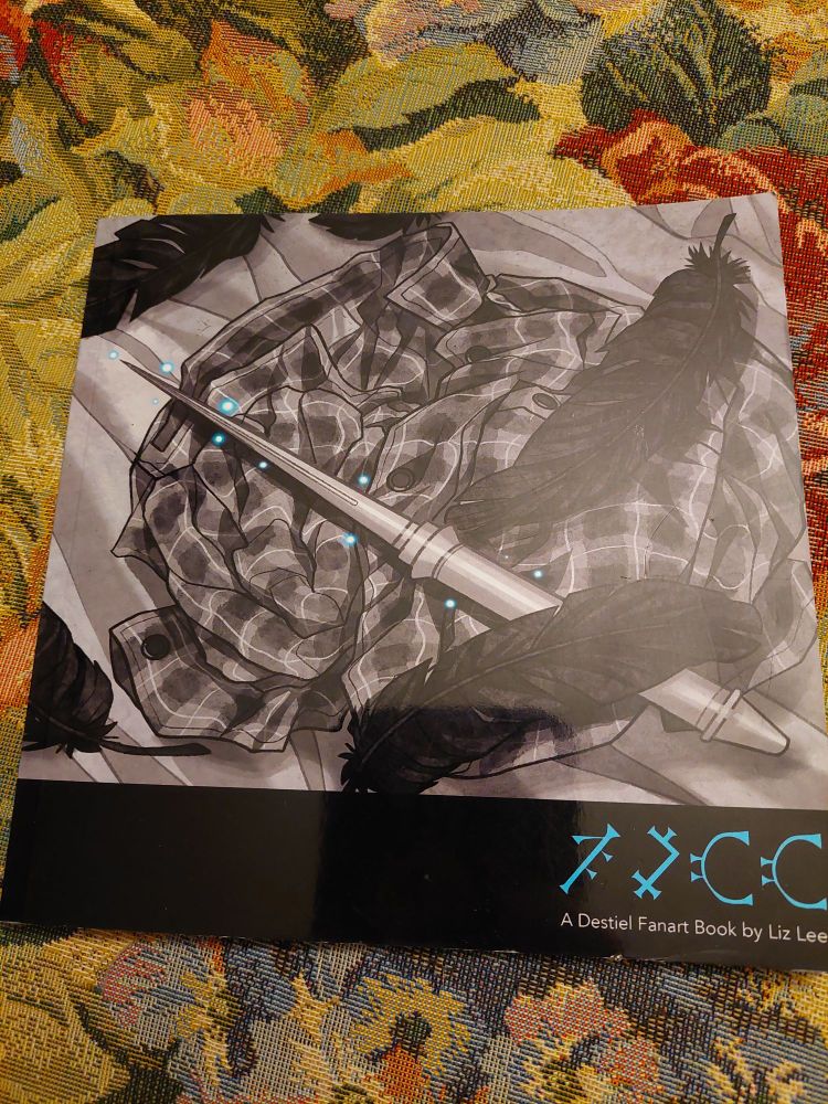 Front cover of a small glossy paperback book. The cover art is of a flannel shirt lying rumpled up, with an angel blade and black feathers on it. In the lower corner there are some Enochian symbols and the text A Destiel Fanart Book by Liz Lee.
