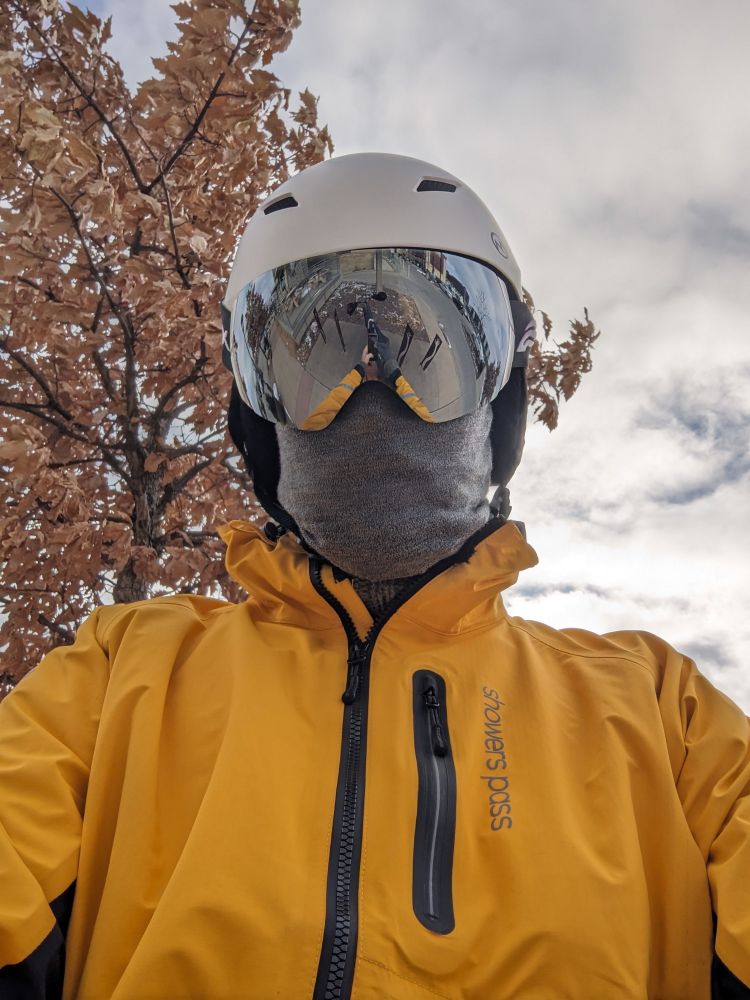 A human clad in a white helmet, reflective goggles, a Grey face mask and a yellow jacket. A tree with tan leaves is in the background. 