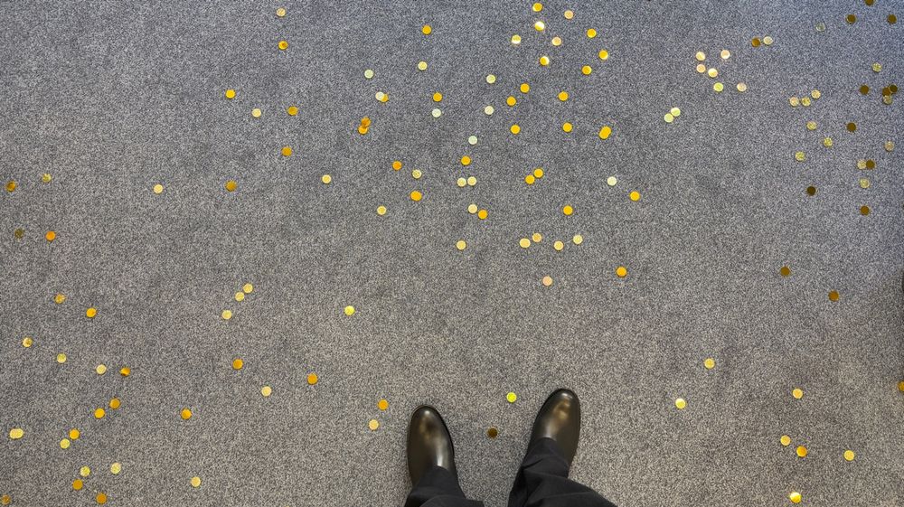 Foto zeigt schwarze Schuhe auf blauem Teppich, der von goldenem Konfetti bedeckt ist. 