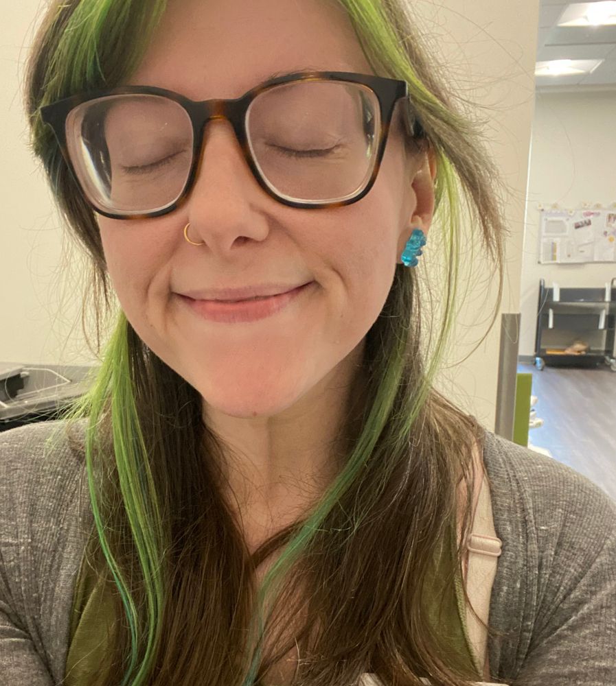 A selfie of Halley, a woman with glasses, a nose ring, and green and brown hair. She is wearing neon blue earrings shaped like gummy bears. 