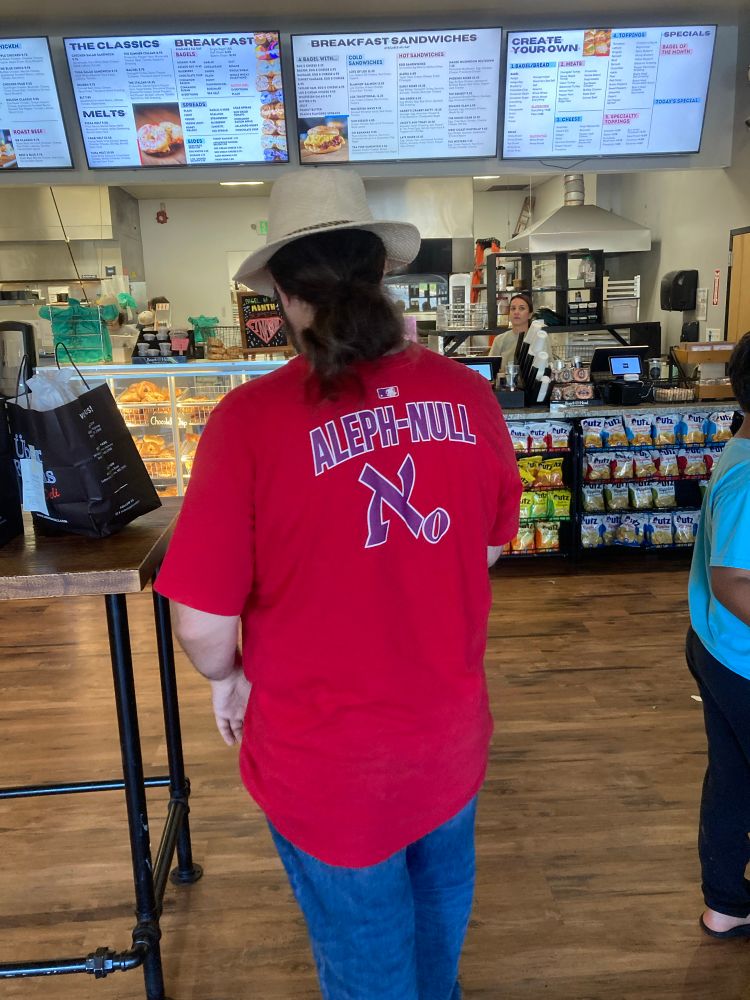 A man in a bagel shop wearing a red baseball jersey with name and number “ALEPH-NULL”
