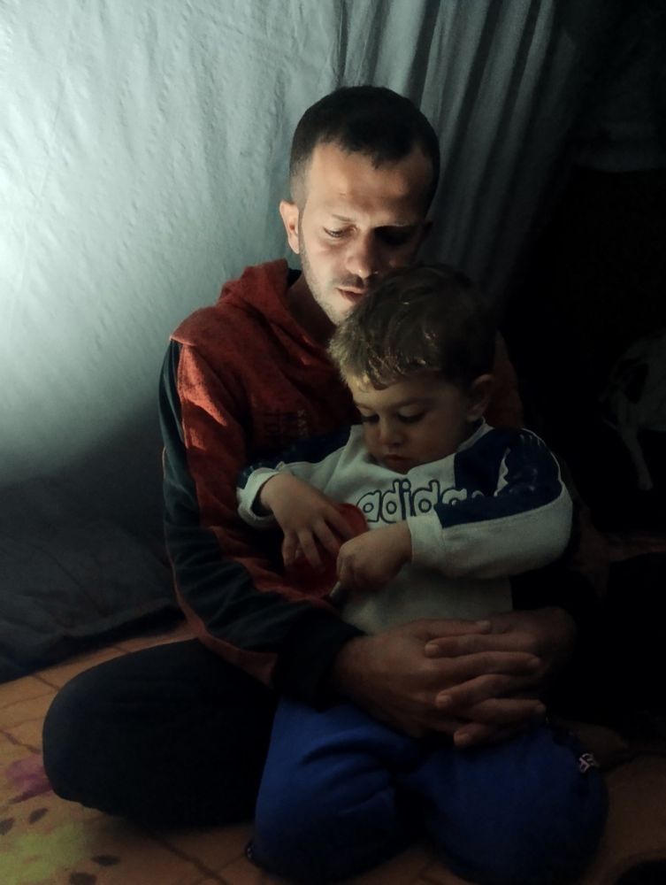 A thin Palestinian man kneeling in a tent with his arms clasped around his young son. They are both dressed for cold weather.