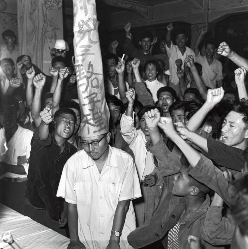 Photo of a Maoist struggle session. A black and white photo of a dejected looking man with glasses is looking by at the floor while a crowd of people with raised fists shout at him 
